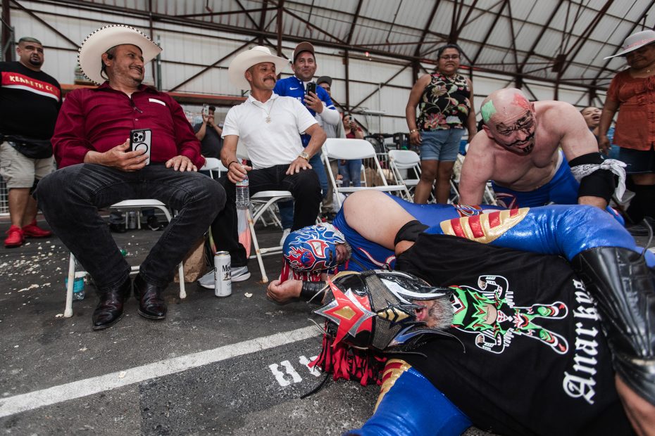 People in a wrestling event, with two wrestlers in colorful masks on the ground in the foreground. Spectators, including two men in cowboy hats, are seated in the background.