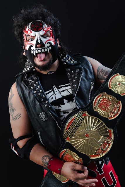 Person wearing face paint, a studded leather vest, and displaying a wrestling championship belt against a black background.