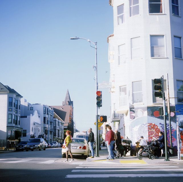 People crossing a street at a crosswalk in an urban area with cars, colorful murals, and a stroller. Weather is clear and sunny.