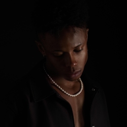 Person wearing a pearl necklace and dark clothing, looking downward against a dark background.