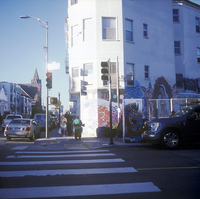 Street scene with a mural on a building corner, pedestrians, cars, and a crosswalk. Bright sunshine casts shadows on the sidewalk.