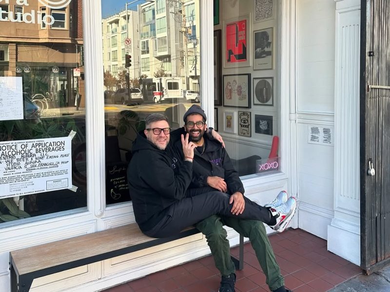 Two people sitting on a bench outside an art studio, with one person sitting on the other's lap. They are smiling, and there are framed artworks displayed in the studio window behind them.