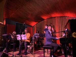 Band performing on stage with a pianist in the foreground, surrounded by musicians playing wind instruments and drums, under warm, red lighting.