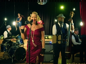 A vintage-style band performs on stage. The lead singer is in a red dress, holding a microphone. Musicians play drums, double bass, and keyboard, with a dancer in the background.