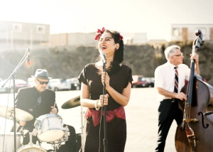 A woman sings into a microphone while a man plays the upright bass and another plays the drums, performing outdoors in a sunlit area.