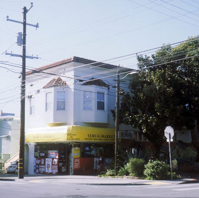 Street corner with a two-story building. The ground floor is a market with a yellow awning and signs. Large tree and power lines are visible.
