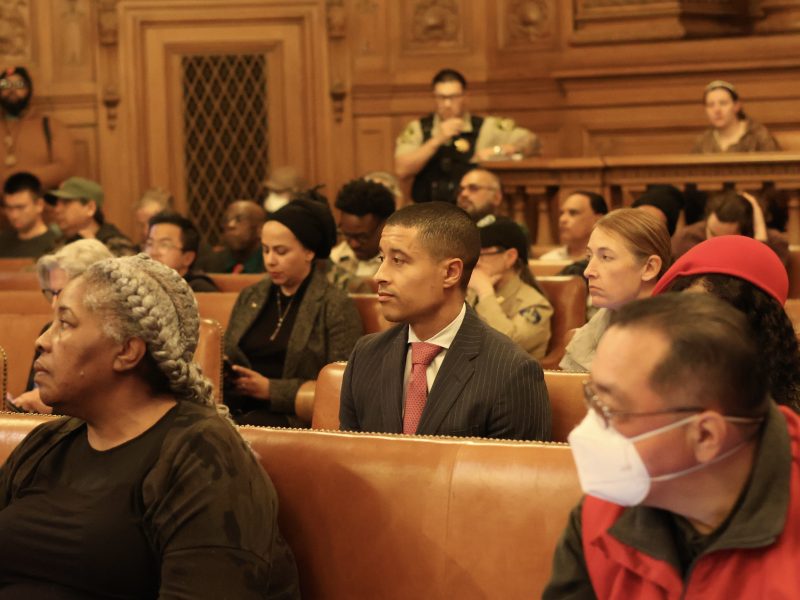 People seated in a wood-paneled courtroom, some wearing masks, with a security guard standing at the back. In one corner, Carter seemed deep in thought amidst the proceedings.