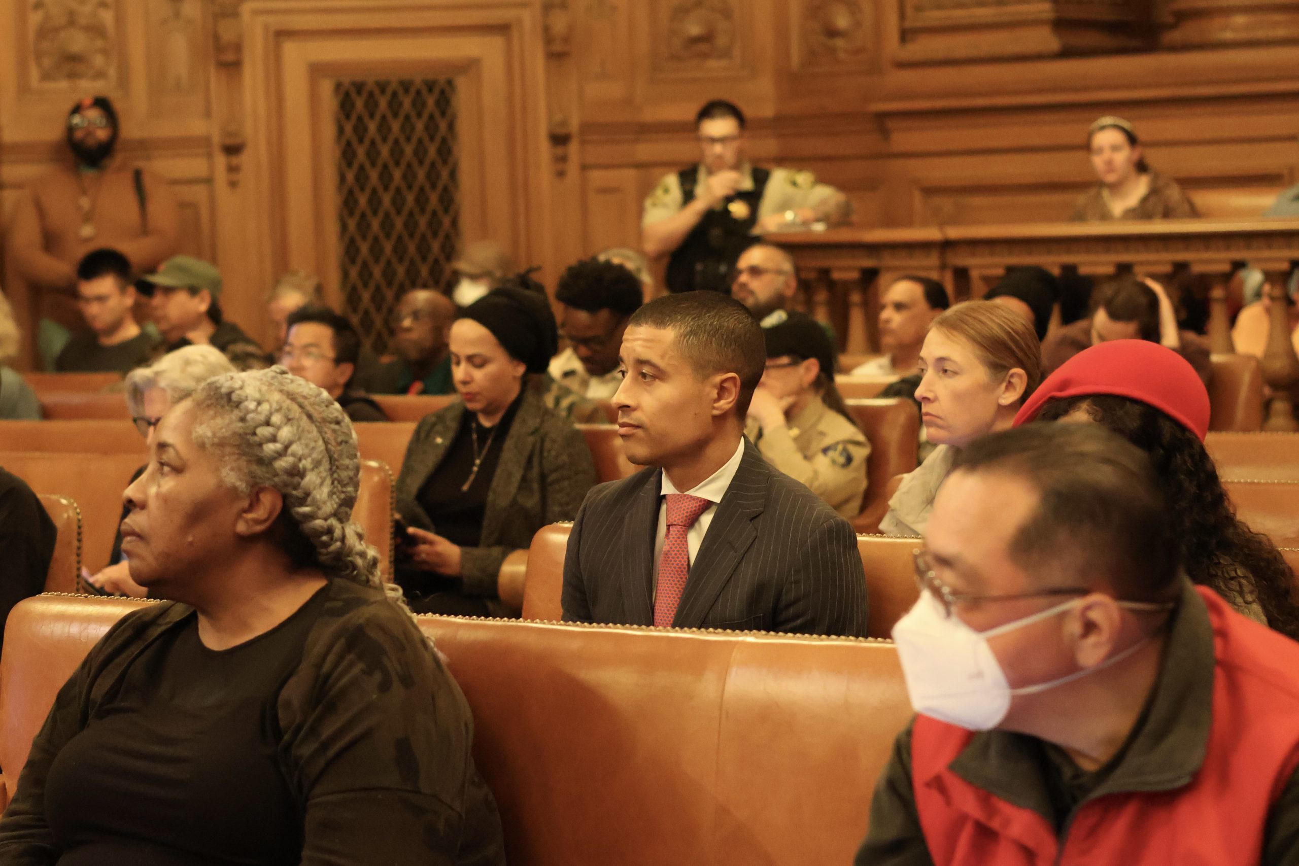 People seated in a wood-paneled courtroom, some wearing masks, with a security guard standing at the back. In one corner, Carter seemed deep in thought amidst the proceedings.