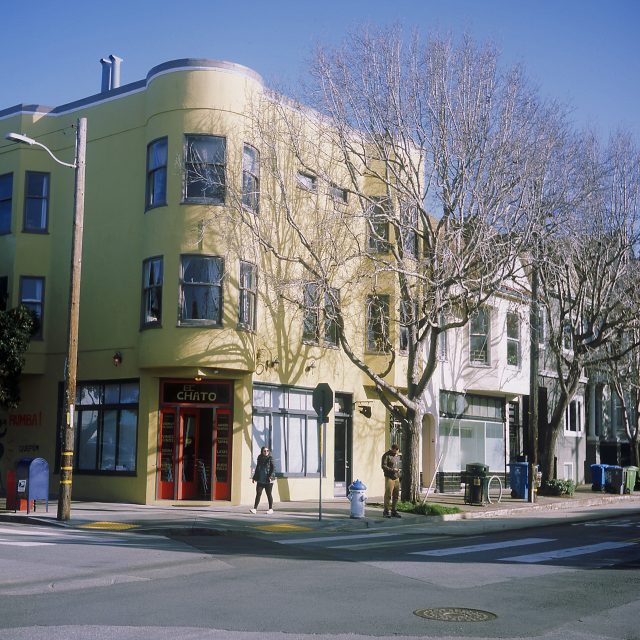 A yellow building with a red door on a street corner. Two people stand outside. Leafless trees line the sidewalk.