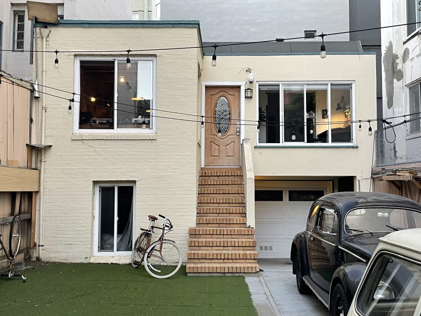 Two-story beige house with a wooden door, large windows, and a garage. A bicycle is parked outside. There's a vintage car in the driveway, with string lights overhead and artificial grass.