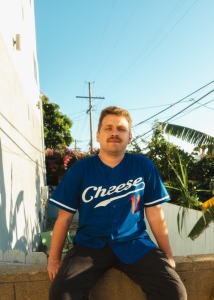 Person with a mustache wearing a blue jersey with "Cheese" text, sitting on a low wall. Background includes a clear sky, power lines, and lush greenery.