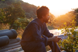 A person in casual clothing sits on a rooftop at sunset, surrounded by greenery, gazing into the distance.