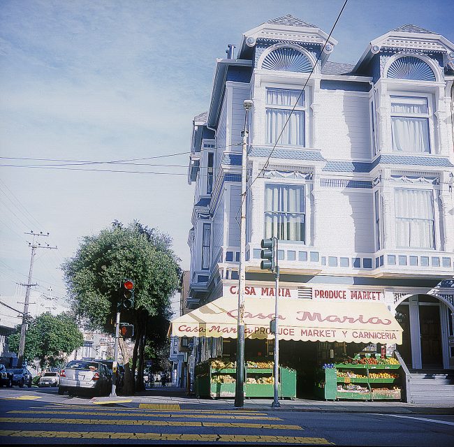 Sunny street corner with a large, ornate building housing "Casa Maria" market below. Produce including oranges and bananas are displayed outside. Traffic lights and a tree are visible.