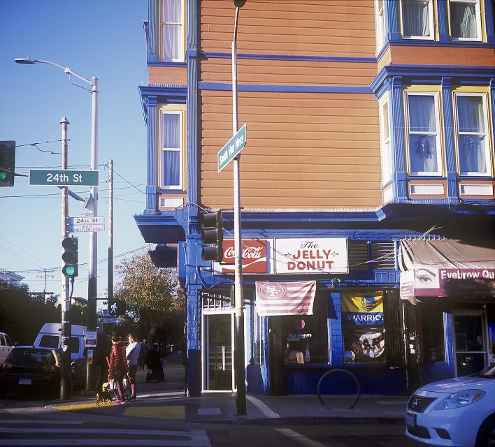 Street corner with a building featuring "The Jelly Donut" shop. People stand near the crosswalk, a car is parked on the street, and street signs note 24th St and South Van Ness Ave.