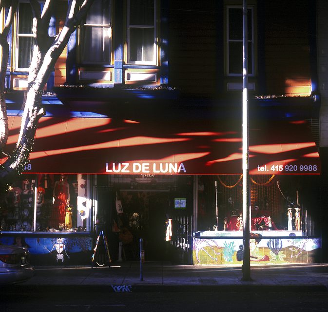 Shopfront with a red "Luz de Luna" awning, flanked by display windows. Bright sunlight casts shadows from trees onto the building.