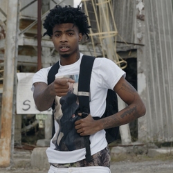 Man wearing a white t-shirt and backpack points forward, standing in front of a dilapidated building.