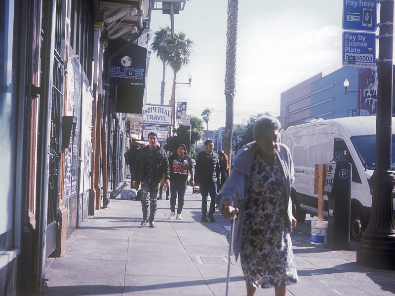 People walking on a sunny sidewalk, with one elderly woman in the foreground using a cane. Buildings, storefronts, and a van are visible in the background.