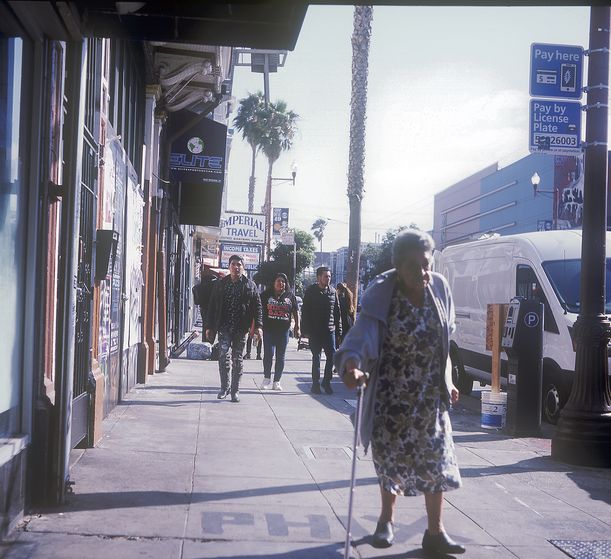 People walking on a sunny sidewalk, with one elderly woman in the foreground using a cane. Buildings, storefronts, and a van are visible in the background.
