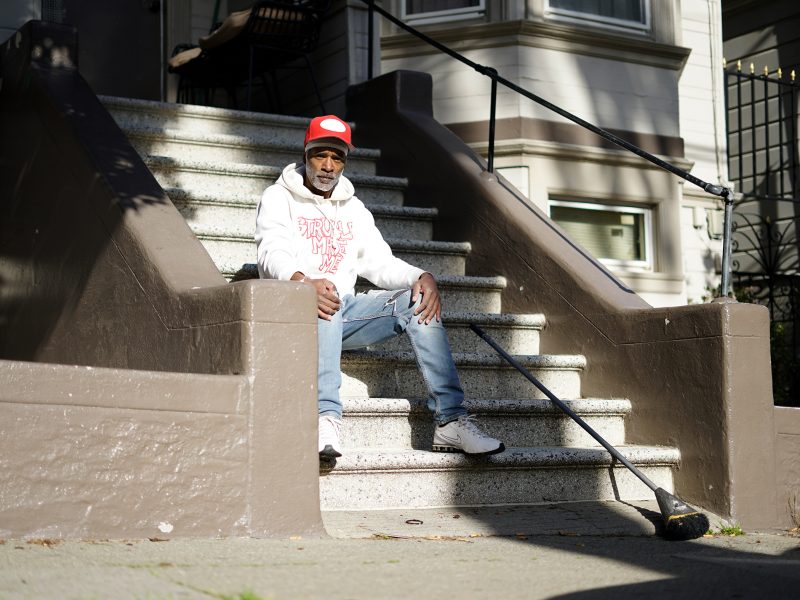 Man in a white hoodie and red cap sits on outdoor steps holding a long rod. Sunny day, building facade in the background.