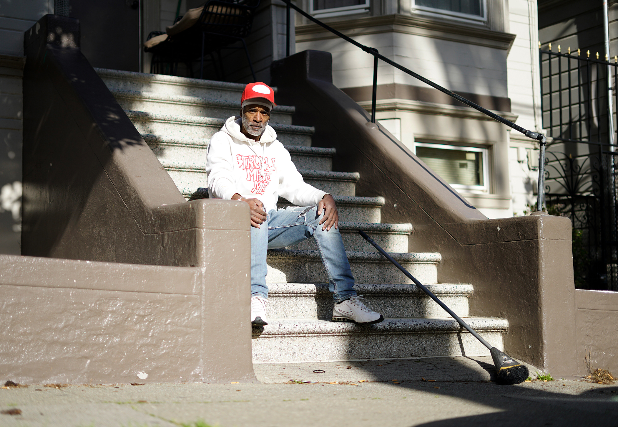 Man in a white hoodie and red cap sits on outdoor steps holding a long rod. Sunny day, building facade in the background.