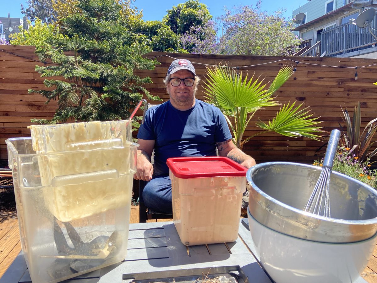 Person sitting outdoors with containers of dough and kitchen tools on a table.
