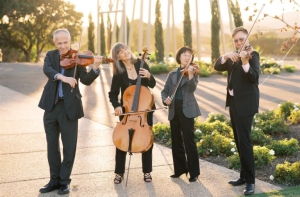 Four musicians outdoors, playing string instruments: two violins, a cello, and a viola. They stand on a paved path surrounded by greenery, with sunlight in the background.