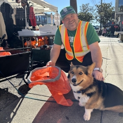 Person in a green shirt and safety vest kneels beside a corgi holding an orange trash bag. They are on a sidewalk with booths and trees in the background.