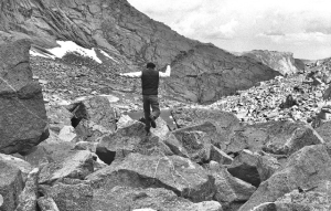 Person standing on large rocks in a mountainous area, with rugged terrain and cloudy sky.
