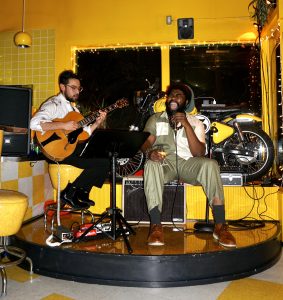 Two men perform on a small elevated stage; one plays guitar, the other sings into a microphone. Behind them is a display with a motorcycle and twinkling lights.