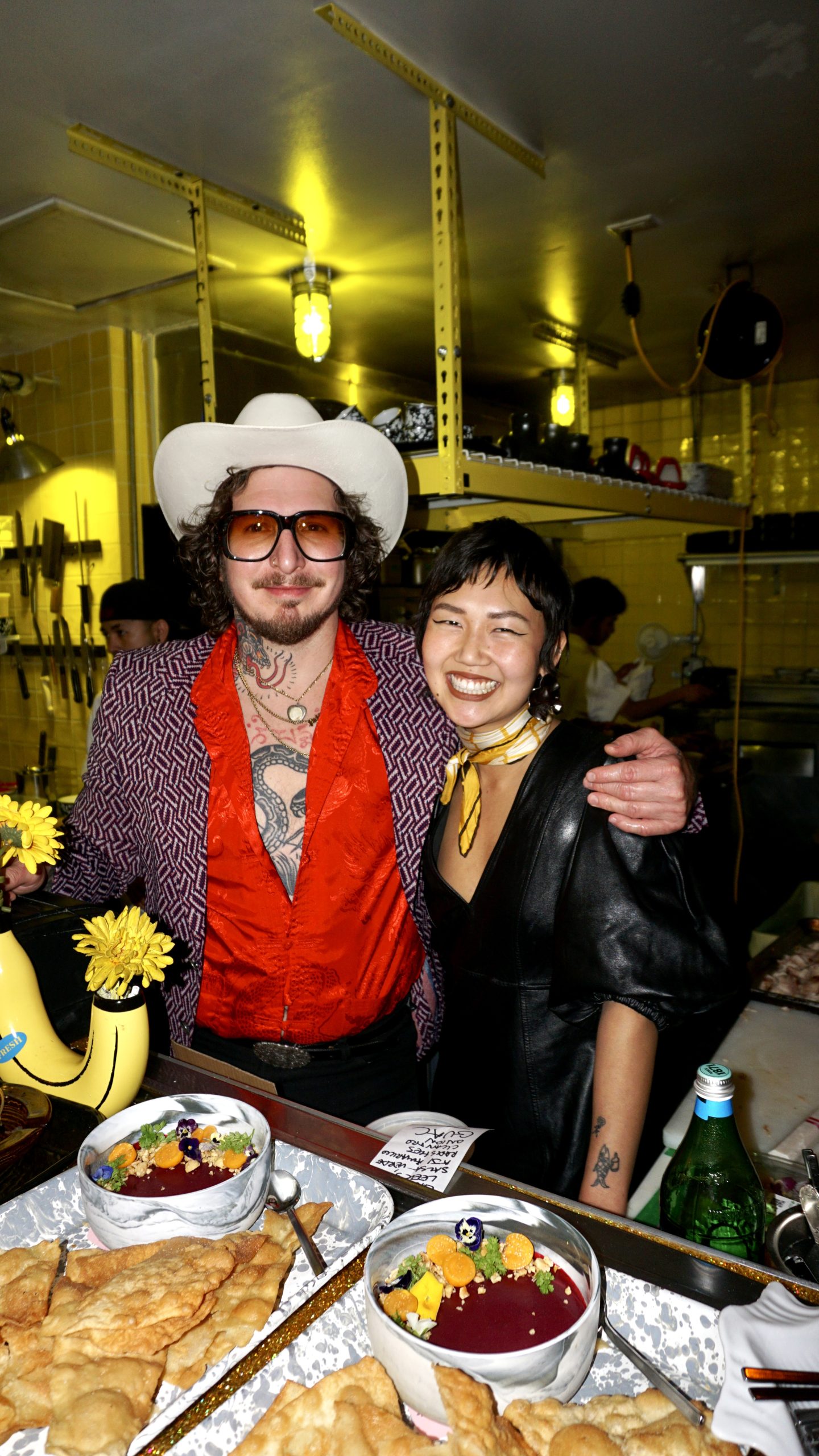 Two people smiling in a kitchen, surrounded by dishes and drinks, with bright yellow lighting and kitchen utensils in the background.