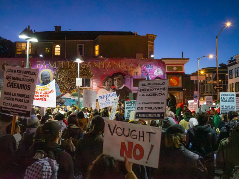 A large crowd gathers for a protest in an urban area. People hold various signs in different languages against political policies. Murals and buildings are visible in the background.