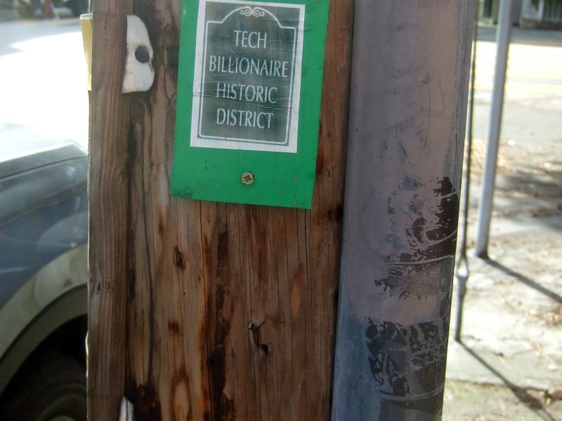 A small green and white sign on a wooden pole reads "Tech Billionaire Historic District."