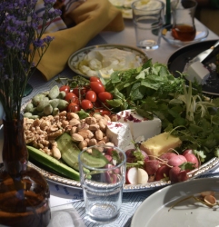 Plate of assorted vegetables, nuts, cheese, and herbs on a table next to a brown vase with purple flowers and a clear glass.
