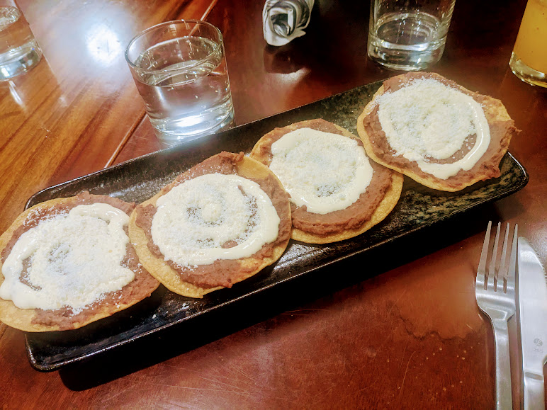 A rectangular black plate with four tostadas topped with refried beans, cream, and cheese. Glasses of water and silverware are on the wooden table.