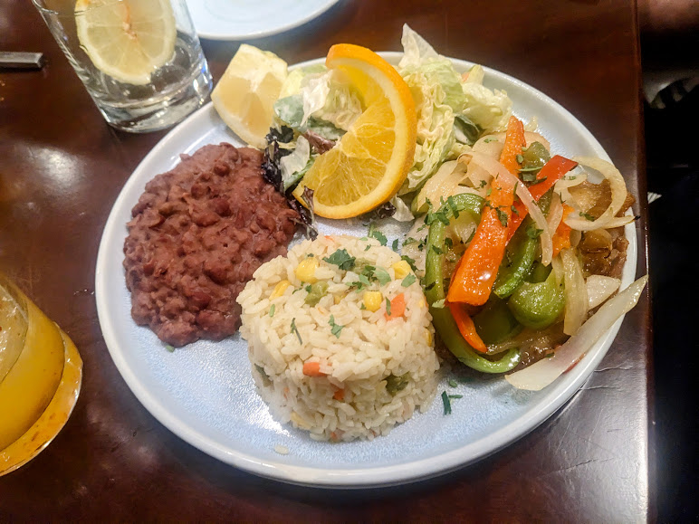 A plate of rice with vegetables, beans, sautéed peppers and onions, and a salad with orange slices, served with lemon wedges.