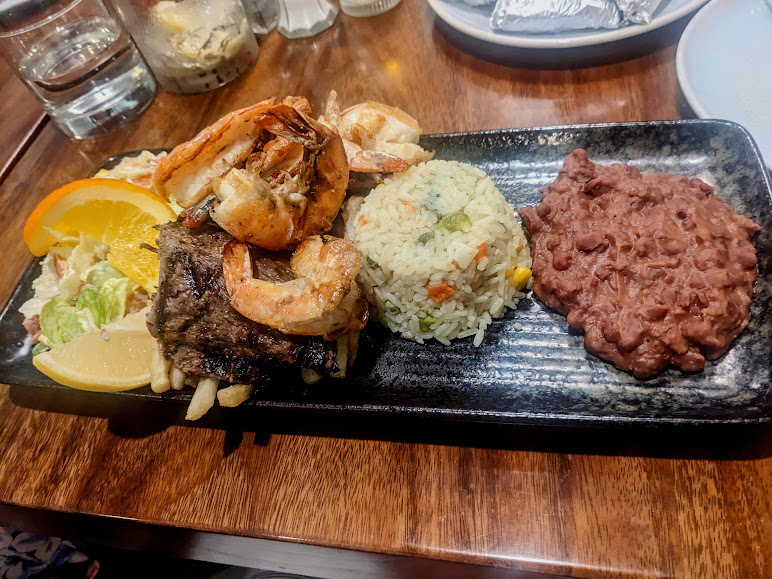 A rectangular plate served with grilled shrimp, beef, rice with vegetables, refried beans, and a mixed salad garnished with lemon and orange slices.