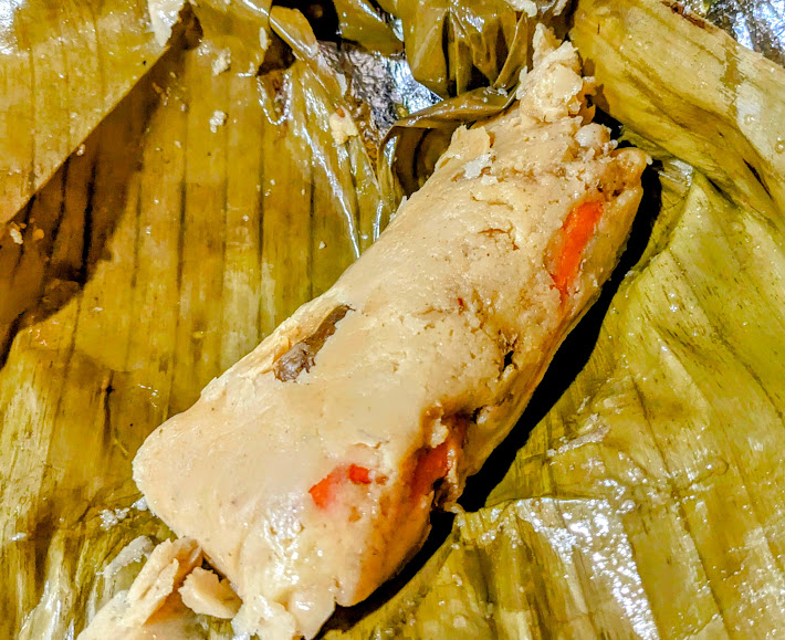 A tamale wrapped in a banana leaf is opened to reveal a soft corn dough with red pepper pieces inside.