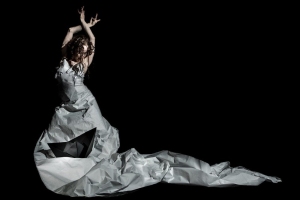 Person in a long, flowing paper dress strikes an expressive pose against a black background.
