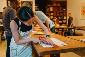 Person in a white apron filleting a fish on a wooden table in a cooking class setting. Other participants and kitchen utensils are visible in the background.