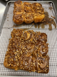 Two trays of glazed cinnamon rolls topped with chopped nuts cooling on wire racks.