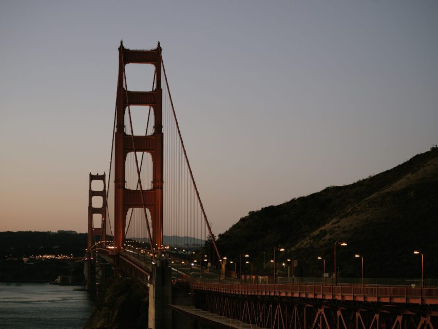 The Golden Gate Bridge at dusk, with its red-orange towers and suspensions, spanning across the water, hills in the background.