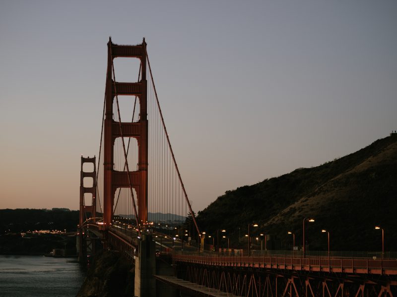 Golden Gate Bridge at dusk with lit streetlamps, spanning a body of water and leading to a hill.