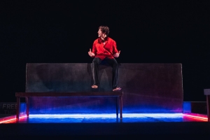 A person in a red shirt and dark pants sits on a raised platform with a dark background, lit by blue and red neon lights underneath.