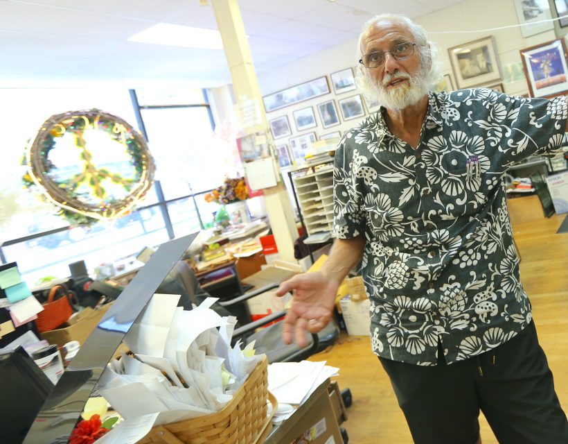 An older man with a white beard wearing a floral shirt stands in a cluttered office, gesturing with his hands. A basket of papers and a wreath are visible in the background.