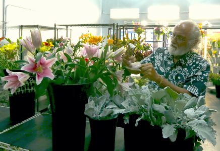 A person in a floral-patterned shirt arranges a bouquet among various flowers and foliage in a dimly lit room.