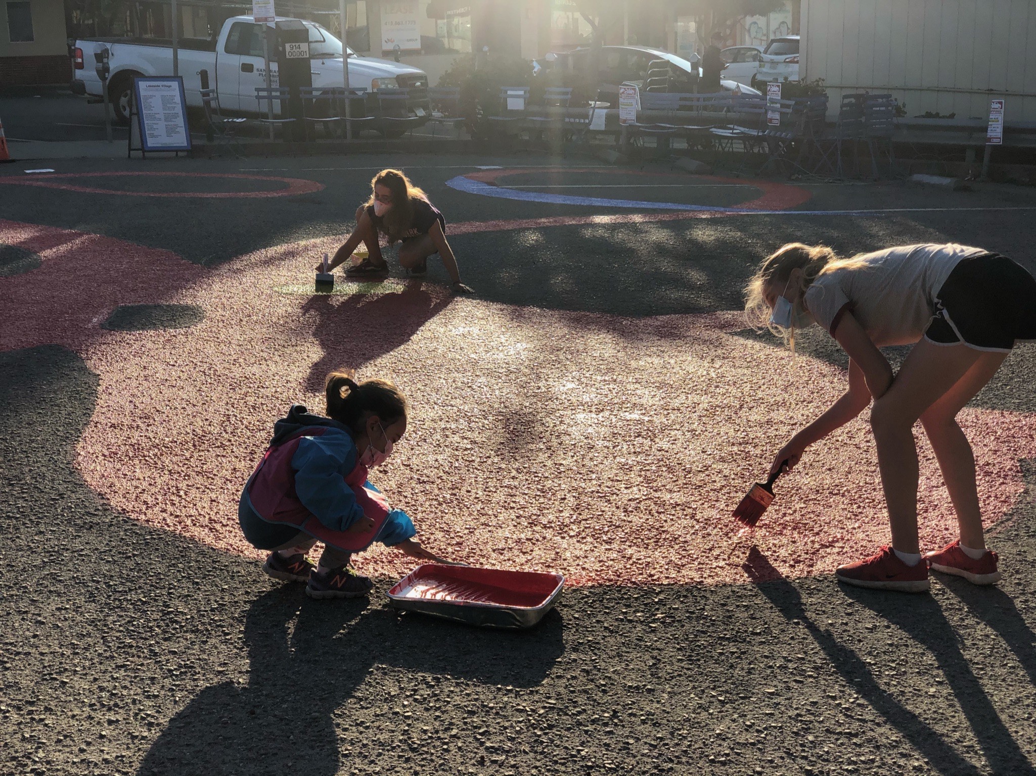 Three people paint a large outdoor mural on the ground in a sunlit area, using paint rollers and brushes.