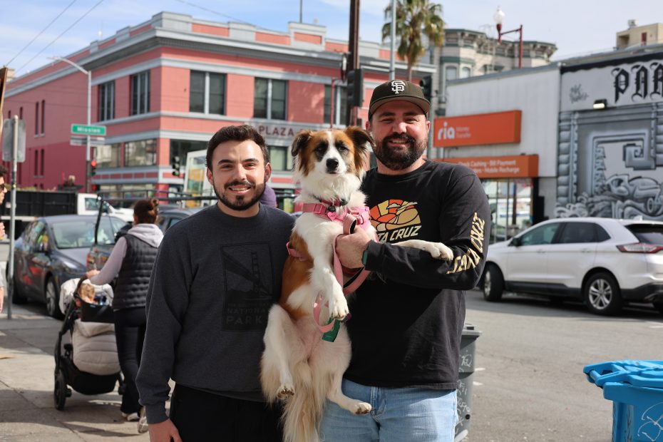 Two men stand smiling on a city street, one holding a dog. There's a red building in the background and some pedestrians.