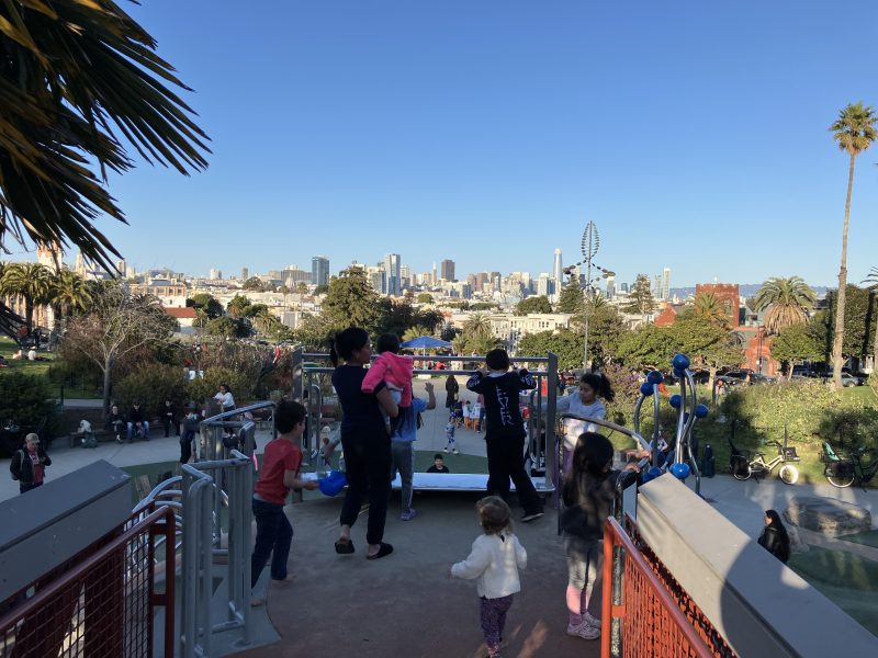 People enjoying a park on a sunny day, with a view of a city skyline in the background.