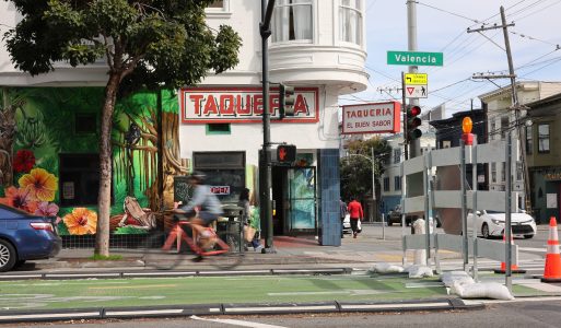 Street view of a taqueria with colorful murals, a cyclist riding past, and a road construction barrier. Signs include "Valencia" and "Taqueria El Buen Sabor.