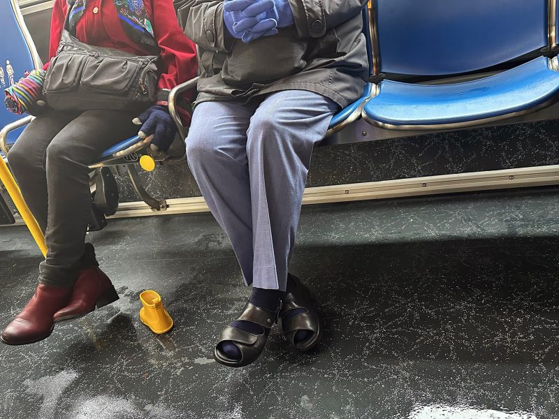 Two people sit on a bus; one is wearing red boots, and the other is in black sandals with blue pants. A yellow rubber boot is on the floor.